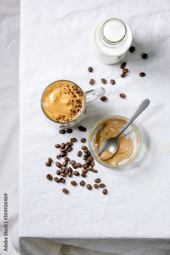 Dalgona frothy coffee trend korean drink milk latte with coffee foam in glass mug, decorated by ground coffee on white cotton table cloth. Ingredients above. Flat lay, space Dalgona frothy coffee trend korean drink milk latte with coffee foam in glass mug, decorated by ground coffee on white cotton table cloth. Ingredients above. Flat lay, space