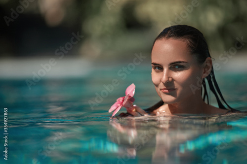 Portrait of brunette stunning lady with wet hair swimming in pool and keeping exotic purple flower in hands. Concept of travelling, relaxing and pleasure