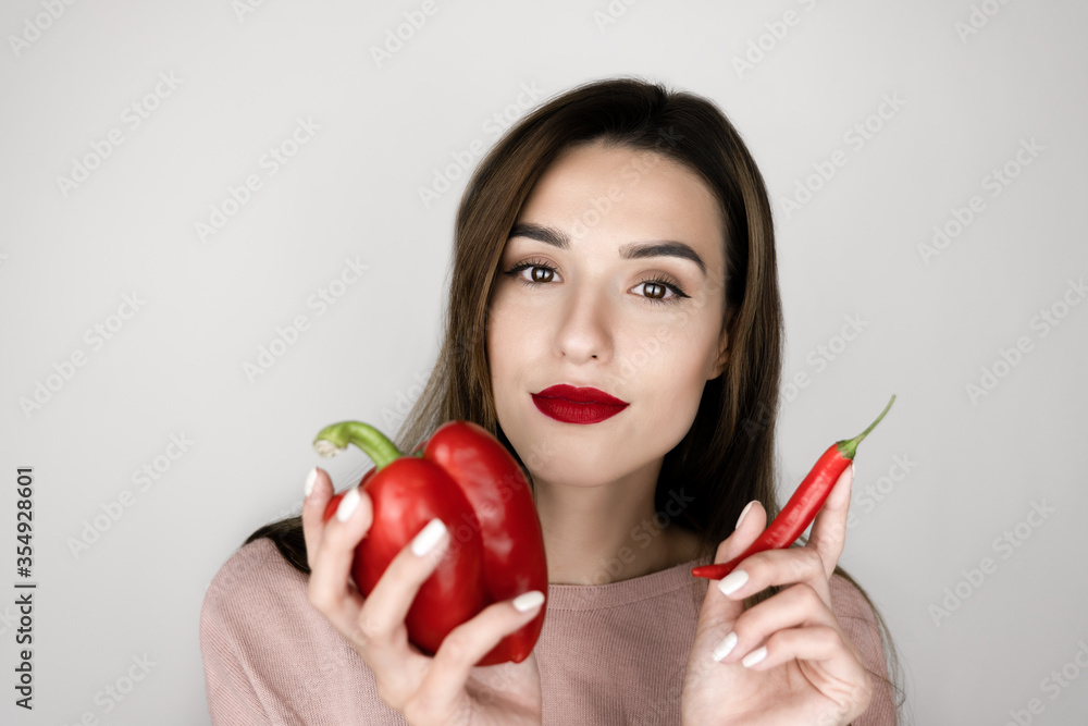 portrait of young brunette woman with red lipstick holding chilly pepper on one hand and sweet red pepper in another , funny mood, isolated white background portrait of young brunette woman with red lipstick holding chilly pepper on one hand and sweet red pepper in another , funny mood, isolated white background