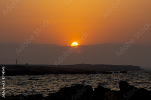Half of yellow sun visible as the sun sets behind the mountains with a rocky shore and sea in the shadows below the beautiful orange sky