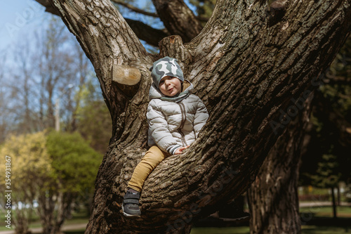 A cute little boy in a camouflage beanie is sitting on the branch of big tree. He is happy to be so high. Image with selective focus © Yulia Raneva