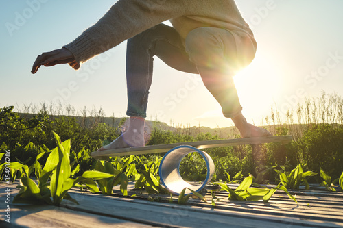 side view of healthy beautiful active woman keeping balance on the wooden balance board against the sky background at sunset summer day. Exercise and outdoor concept.