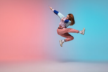 In jump. Beautiful girl dancing hip-hop in stylish clothes on colorful gradient background at dance hall in neon light. Youth culture, movement, style and fashion, action. Fashionable bright portrait.