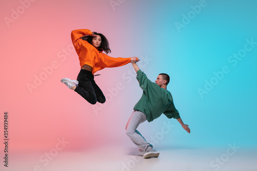 Fototapeta Naklejka Na Ścianę i Meble -  In jump. Boy and girl dancing hip-hop in stylish clothes on colorful gradient background at dance hall in neon. Youth culture, movement, style and fashion, action. Fashionable portrait. Street dance.