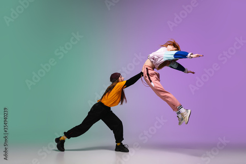Beautiful sportive girls dancing hip-hop in stylish clothes on colorful gradient background at dance hall in neon light. Youth culture, movement, style and fashion, action. Fashionable bright portrait