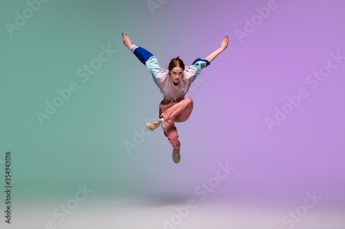 In jump. Beautiful girl dancing hip-hop in stylish clothes on colorful gradient background at dance hall in neon light. Youth culture, movement, style and fashion, action. Fashionable bright portrait.
