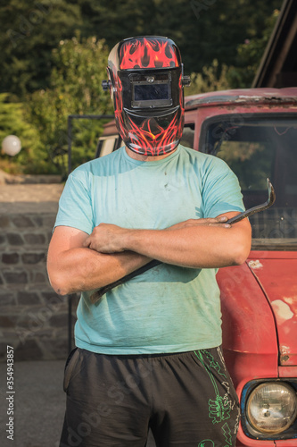 Man with a welding mask on his head in front of a vintage rusty and destroyed truck or van. Car restoration in progress. Tough guy restoring a car