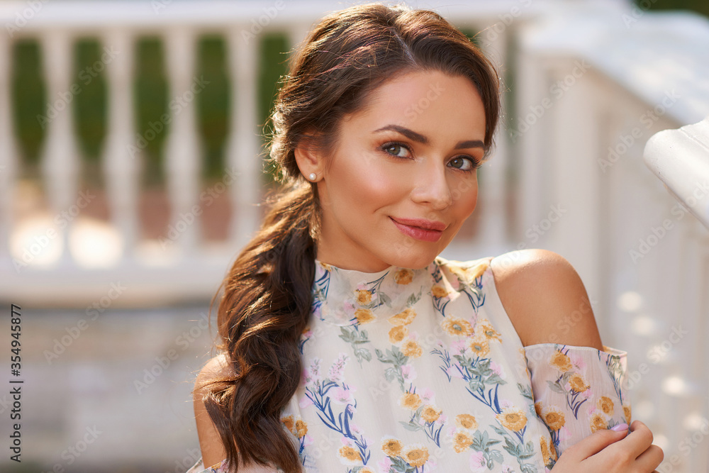 Pretty smiling young woman with long brunette hair wearing fashionable floral dress standing on street against balustrade on background. Happy female model in elegant stylish outfit posing outdoors.
