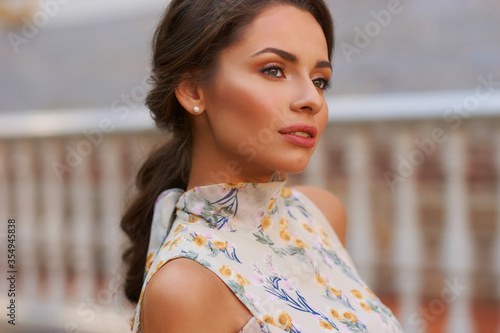 Obraz na plátně Pretty smiling young woman with long brunette hair wearing fashionable floral dress standing on street against balustrade on background