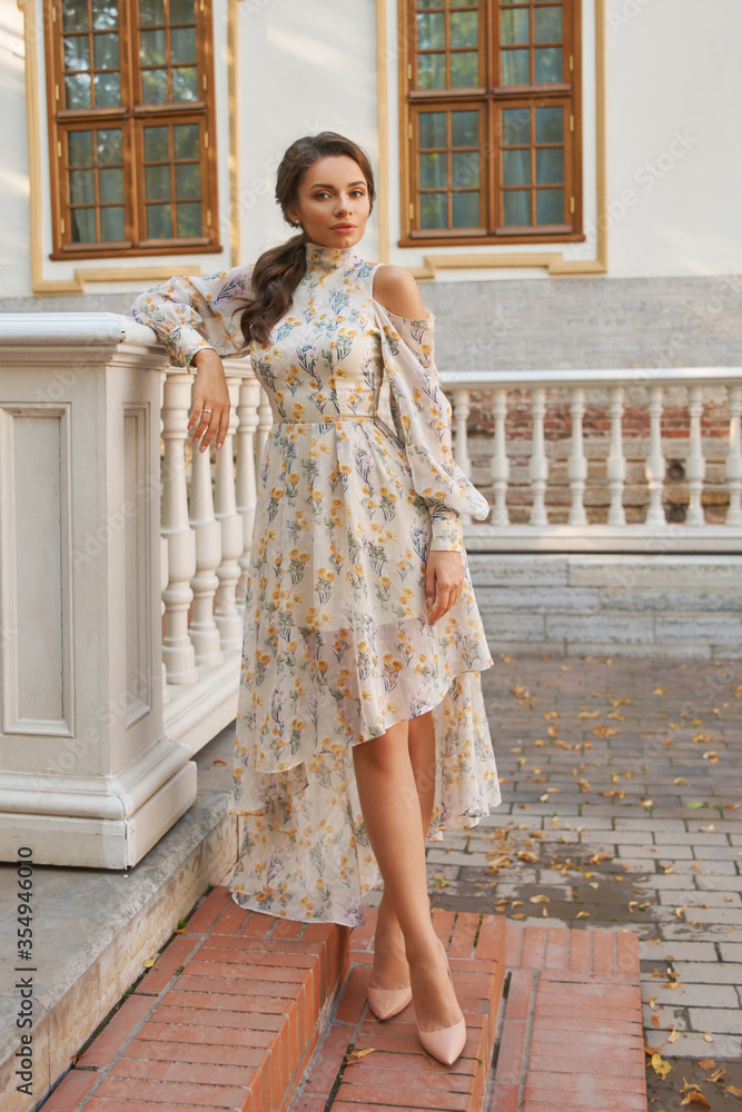 Full-body portrait of young woman wearing elegant chiffon floral dress standing at street and leaning on balustrade. Gorgeous brunette model in summer outfit posing outside exquisite building.