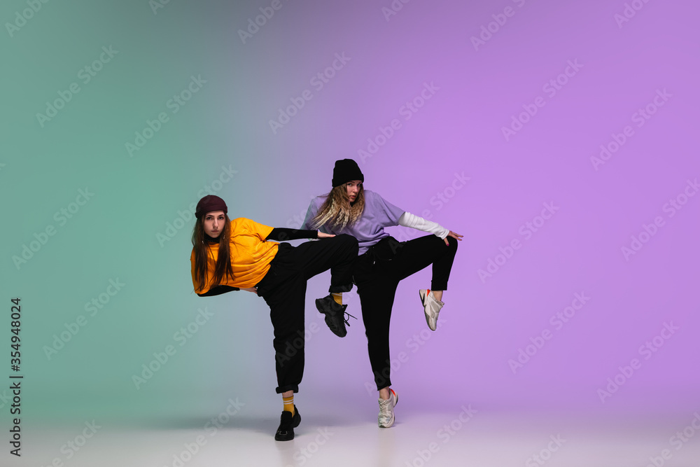 Beautiful sportive girls dancing hip-hop in stylish clothes on colorful gradient background at dance hall in neon light. Youth culture, movement, style and fashion, action. Fashionable bright portrait
