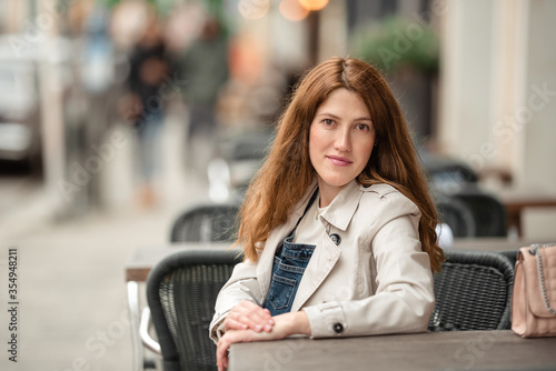 Beautiful young pregnant woman wearing sweater, jeans, grey coat sitting outside a cafe with a beautiful exterior in the summer.