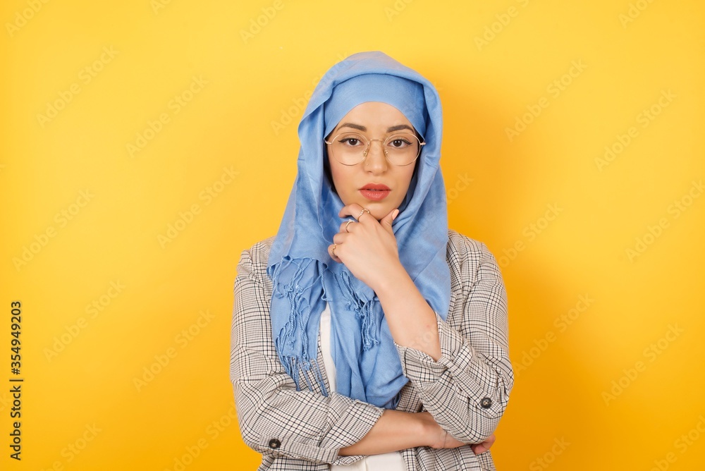 Portrait of thoughtful smiling muslim girl keeps hand under chin, looks directly at camera, listens something with interest, dressed casually, poses against studio wall. Youth concept.