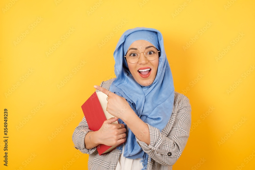 Look over there! Young beautiful muslim woman wearing hijab pointing away and smiling to you while standing against studio background. Look over there! Young beautiful muslim woman wearing hijab pointing away and smiling to you while standing against studio background.