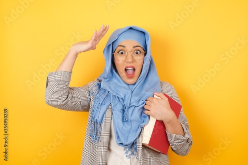 Beautiful young muslim woman wearing hijab female looks with excitement at camera, keeps hands raised over head, notices something unexpected, isolated over wall. Lovely woman reacts on sudden news. © Jihan