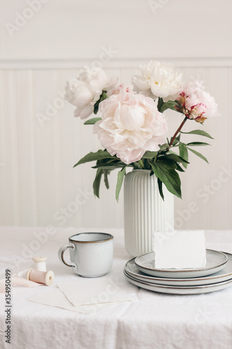 Wedding still life scene. Business, place card mockup scene, marble tray, cup of coffee an pink peony flowers in vase. White linen table cloth. Floral festive feminine styled photo, vertical.