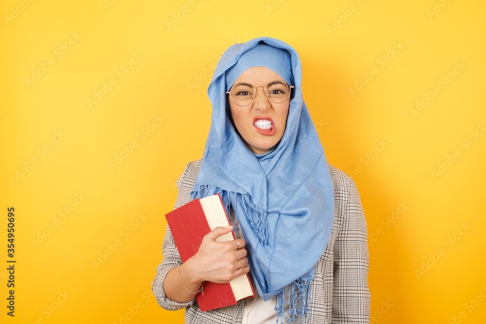Mad crazy young muslim woman clenches teeth angrily, being annoyed with coming noise, dressed in fashionable clothes, isolated over studio background. Negative feeling concept. Mad crazy young muslim woman clenches teeth angrily, being annoyed with coming noise, dressed in fashionable clothes, isolated over studio background. Negative feeling concept.
