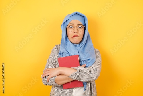 Isolated portrait of stylish young muslim woman wearing hijab bitting her mouth and looking worried and scared crossing arms, worry and doubt. © Jihan