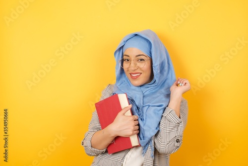 Image of cheerful pretty young muslim woman standing against studio wall with hand near face. Looking with glad expression at the camera after listening to good news. Confident girl. © Jihan