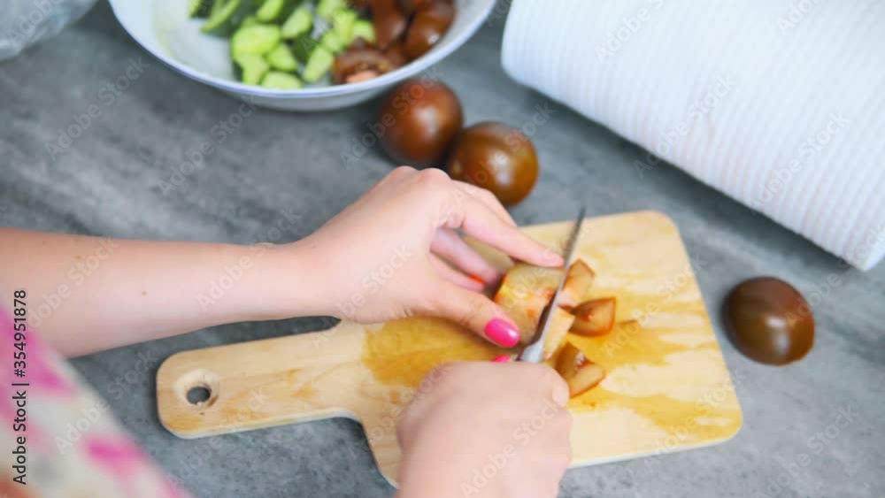 Woman is cutting kumato tomatoes on a wooden board. A bowl with cut cucumbers and tomatoes is on the background. High quality 4k footage