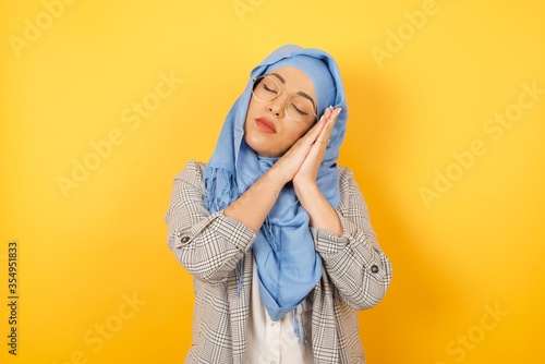 Relax and sleep time. Tired beautiful young muslim woman wearing hijab with closed eyes leaning on palms as pillow pretending sleeping being exhausted, standing against background. Sleeping gesture.