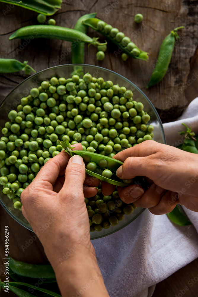 White female hands holding a handful green peas on a wooden table. In the table there are a crystal bowl full of green peas and a linen cloth. Rustic style. Top view. White female hands holding a handful green peas on a wooden table. In the table there are a crystal bowl full of green peas and a linen cloth. Rustic style. Top view.