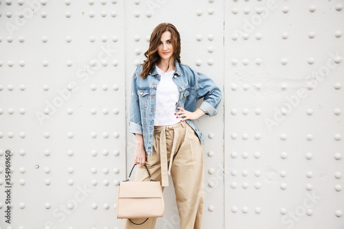 Outdoors fashion portrait of trendy pretty girl posing on the white wall background. Smiling and walking on the city. Going shopping. Wearing stylish wide jean jacket and beige slacks. Pensive mood