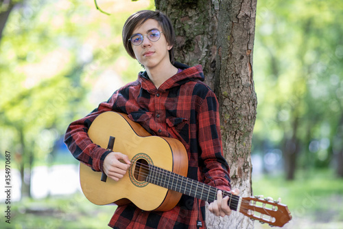 a young man plays an acoustic guitar in a Park leaning against a tree
