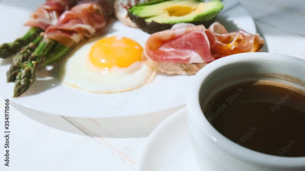 fried asparagus wrapped in jamon, fried eggs, bruschetta with soft cheese and avocado on a white plate and a cup of freshly brewed coffee fried asparagus wrapped in jamon, fried eggs, bruschetta with soft cheese and avocado on a white plate and a cup of freshly brewed coffee