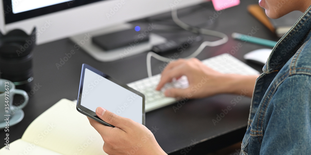A smart man is holding a white blank screen tablet while typing on a keyboard at the black working desk. A smart man is holding a white blank screen tablet while typing on a keyboard at the black working desk.