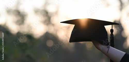Cropped image of university student hand holding a graduation hat in hand over outdoors with sunset as background. photo