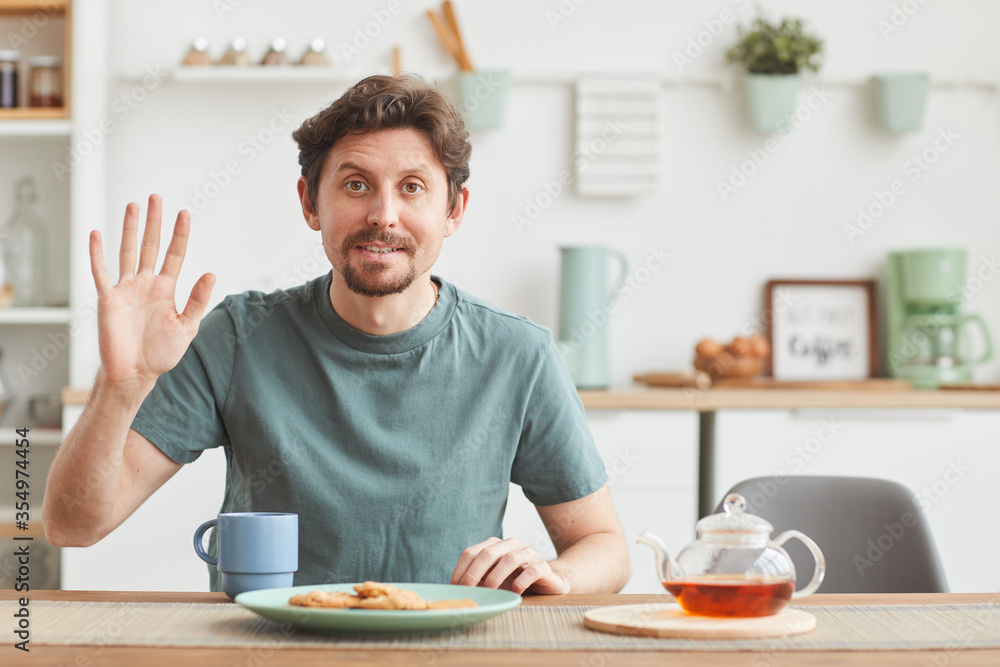 Portrait of young man looking at camera and waving his hand while having breakfast in the kitchen Portrait of young man looking at camera and waving his hand while having breakfast in the kitchen