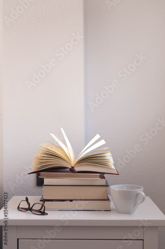 libro abierto sobre varios libros en una mesita de noche blanca, sobre la que tambien hay unas gafas de ver y una taza blanca de desayuno, con una pared blanca al fondo photo