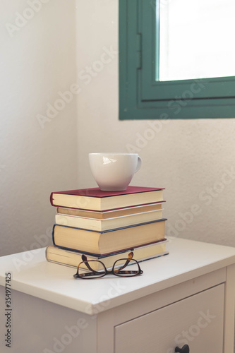 una taza blanca sobre una pila de libros, y unas gafas de ver sobre una mesita de noche blanca, con una ventana al fondo © Rebeca