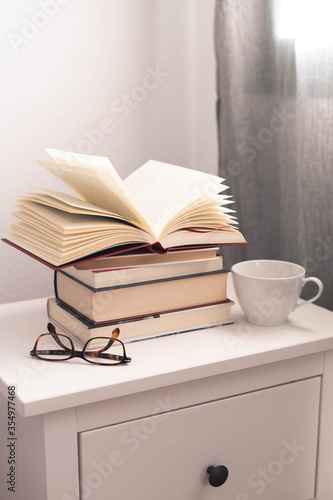 libro abierto sobre una pila de libros en una mesita de noche con una taza blanza y unas gafas de ver photo