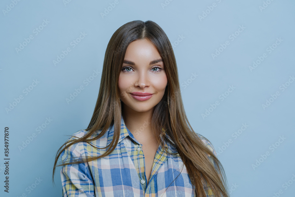 Isolated shot of pretty businesswoman has pleased face expression, tender smile, dressed in casual shirt, has clear skin and attractive facial features, gazes sensually at camera, blue background Isolated shot of pretty businesswoman has pleased face expression, tender smile, dressed in casual shirt, has clear skin and attractive facial features, gazes sensually at camera, blue background