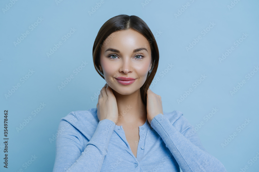 Headshot of young lovely woman keeps hands on neck, has European appearance, happy to hear pleasant words, wears casual sweater, isolated over blue background. Human face expressions concept Headshot of young lovely woman keeps hands on neck, has European appearance, happy to hear pleasant words, wears casual sweater, isolated over blue background. Human face expressions concept