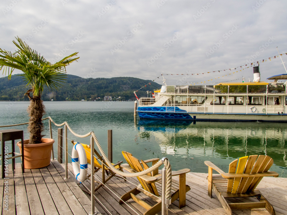 Umbrella, table and chairs placed on deck that borders calm Wörthersee in Austria with ferry on water in the background. 
