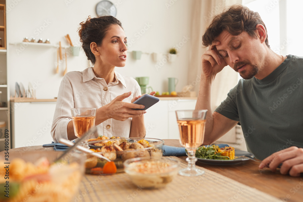 Young woman pointing at mobile phone and talking to man during dinner in the kitchen at home Young woman pointing at mobile phone and talking to man during dinner in the kitchen at home