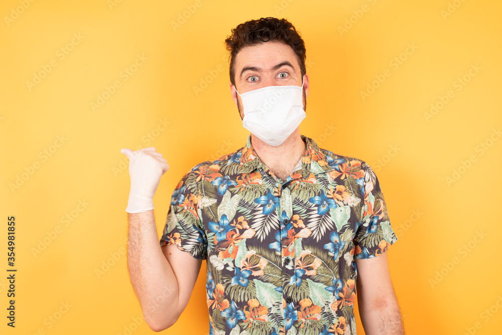 Close up photo of lovely young man pointing aside with forefinger, showing at copy space having news about bargains isolated over yellow background. Wearing Hawaiian shirt and medical mask