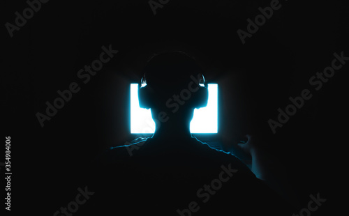silhouette of a human in headphones sitting in front of a bright screen in the dark