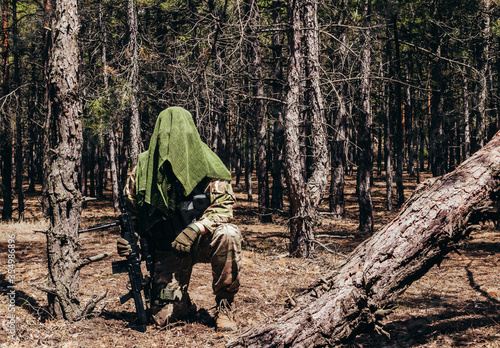 Forest sniper in camouflage and net scarf disguise sitting with rifle in woods.