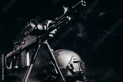 Sniper rifle with scope standing on table with soldier armor helmet on black background with smoke.