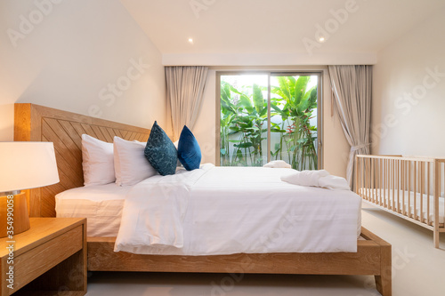 Interior design of bedroom in luxury villa, apartment feature bed, beding, duvet, towels, cushion, bedside table lamp, baby crib, baby cot and sluding door with garden view