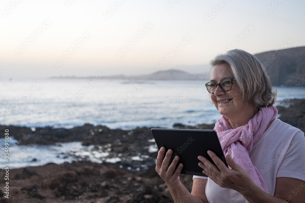 Adult senior woman gray hair and pink scarf sitting near the beach whit the tablet computer. Dusk light. Mountains and sea in background.