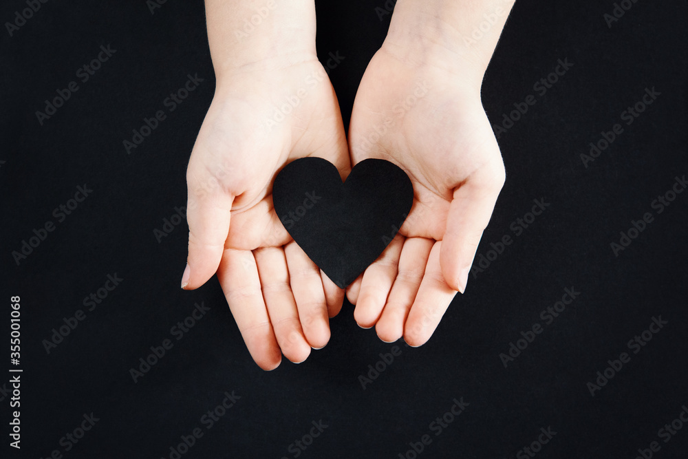 Obraz premium White human hands holding black paper heart on dark background. Person support activist of USA movement black lives matter. People protest against racism. Blackout Tuesday, 2020.