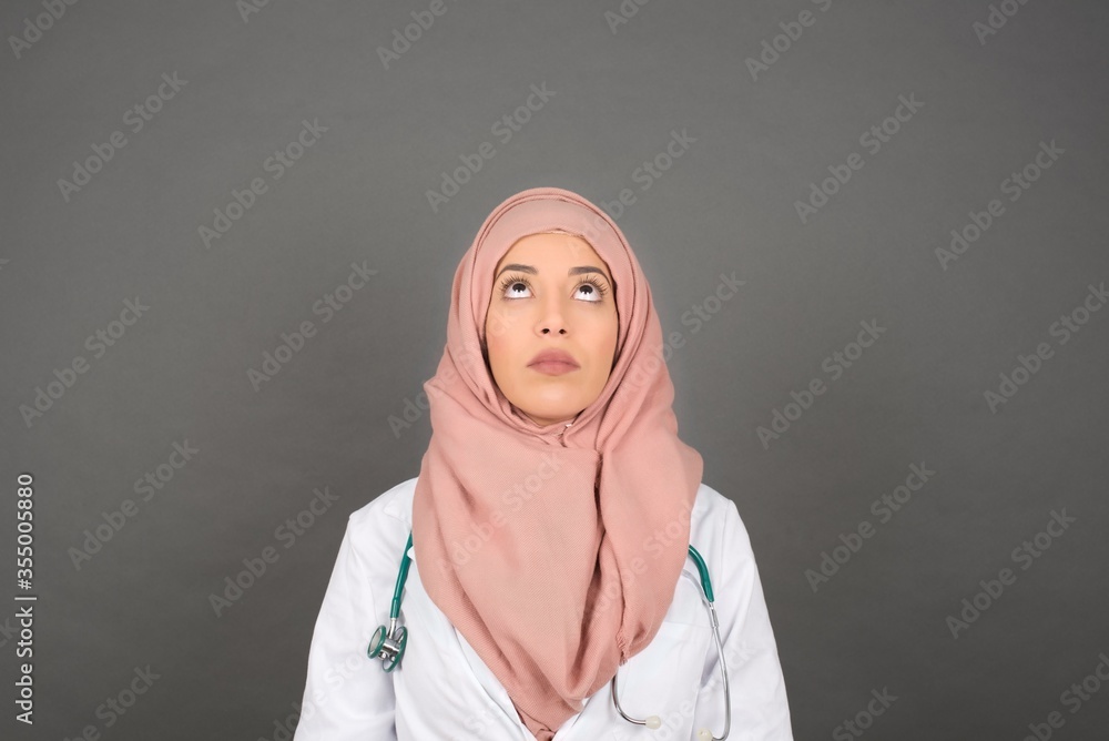 portrait of mysterious muslim doctor woman with straight hair looking up with enigmatic smile. Beautiful smiling girl looking up standing against gray wall. portrait of mysterious muslim doctor woman with straight hair looking up with enigmatic smile. Beautiful smiling girl looking up standing against gray wall.