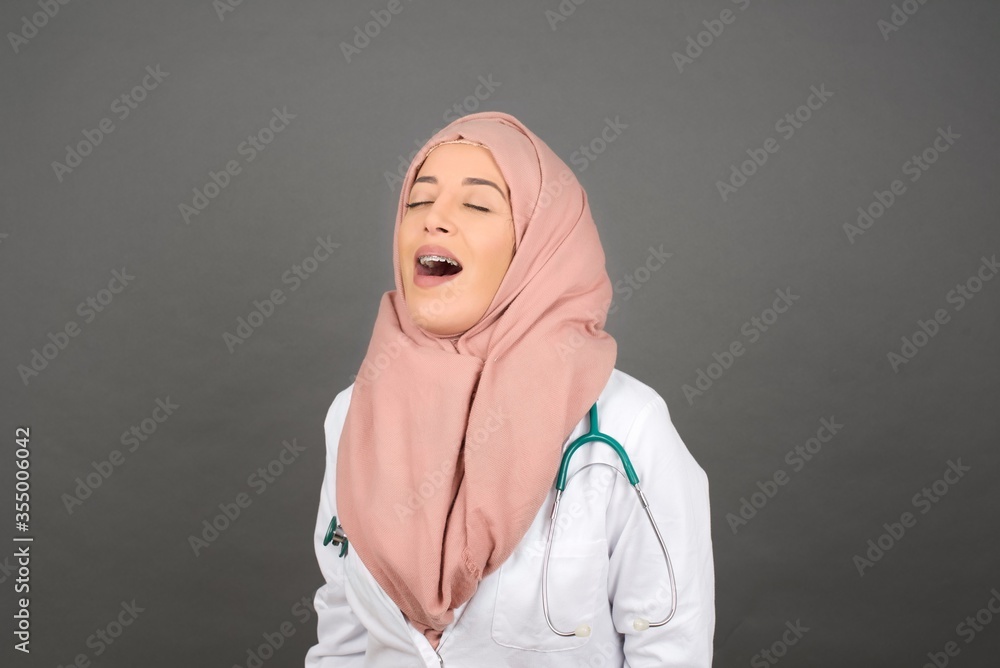 Joyful muslim doctor woman having fun and laughs at good joke or sings wears casual clothes, standing against gray wall. Happy woman with long hair poses inddoors. Joyful muslim doctor woman having fun and laughs at good joke or sings wears casual clothes, standing against gray wall. Happy woman with long hair poses inddoors.