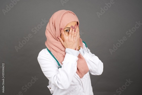 Beautiful young caucasian doctor woman covering her face with her hands and peering out with one eye between her fingers standing indoors. Scared from something or someone.