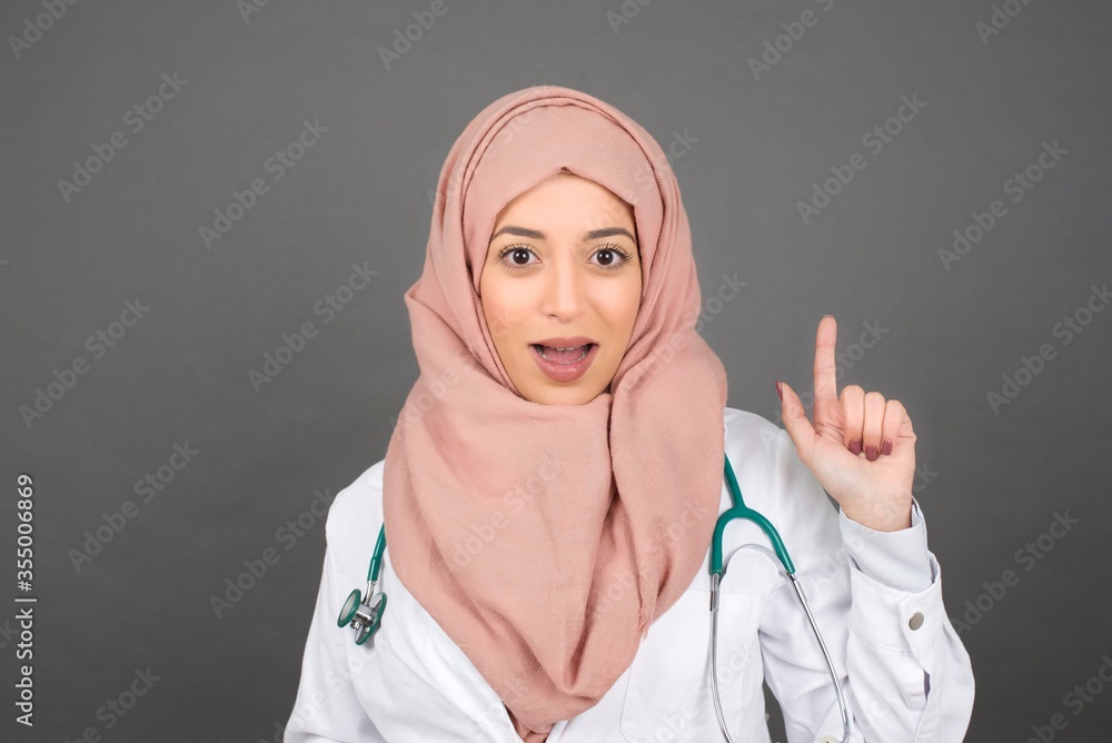 Close up portrait of pleasant looking muslim young doctor woman model has clever expression, raises one finger, remembers herself not to forget tell important thing. Indoors Close up portrait of pleasant looking muslim young doctor woman model has clever expression, raises one finger, remembers herself not to forget tell important thing. Indoors